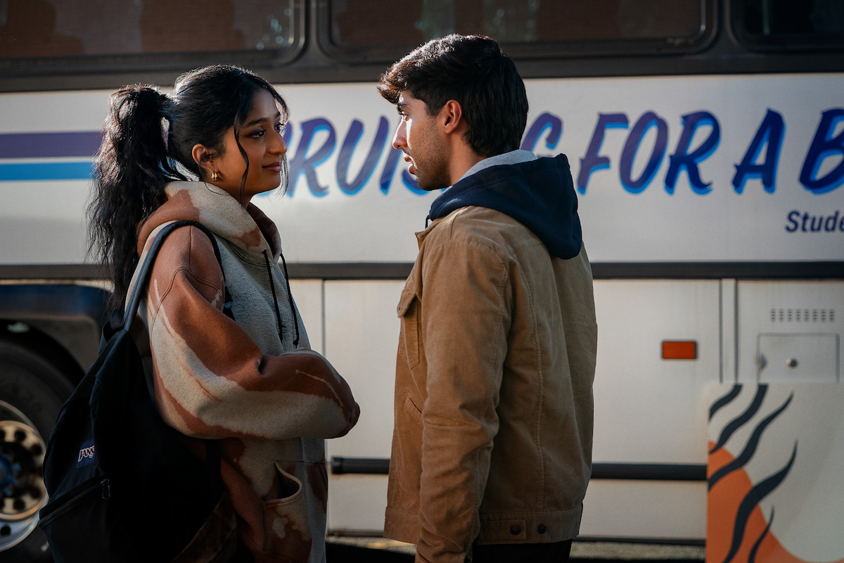 Two young adults stand facing each other at a bus stop, engaged in a serious conversation. There is a large bus with partial text in the background, suggesting a travel or departure scene.