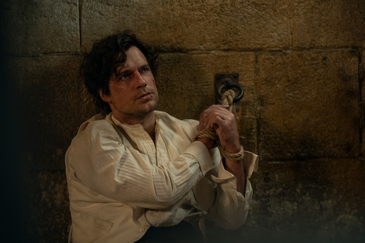 A man in a white shirt sits on the floor of a dim, stone-walled cell, his hands bound with rope and chained to the wall, with a distressed expression on his face.