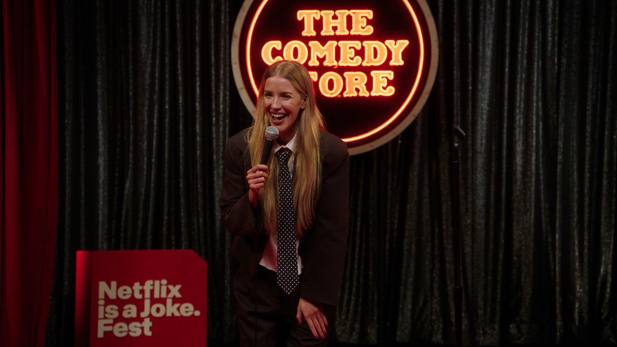 A comedian performs on stage at The Comedy Store, holding a microphone and smiling, with a curtain backdrop and a sign for Netflix is a Joke Fest visible on the left.