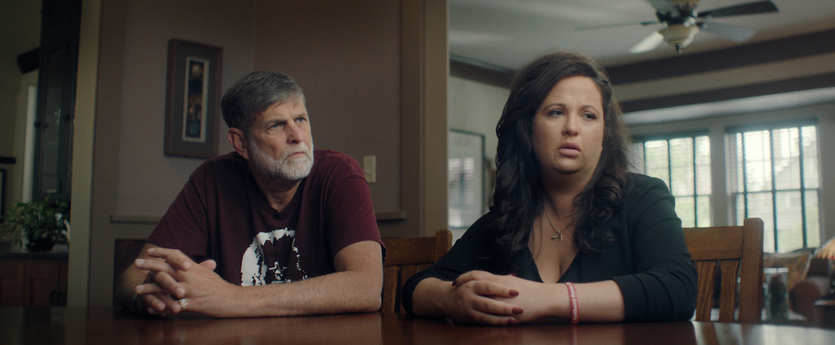 Two people, a man and a woman, sit at a wooden table in a well-lit home dining area with windows, ceiling fan, and framed wall art, both looking thoughtful and serious.