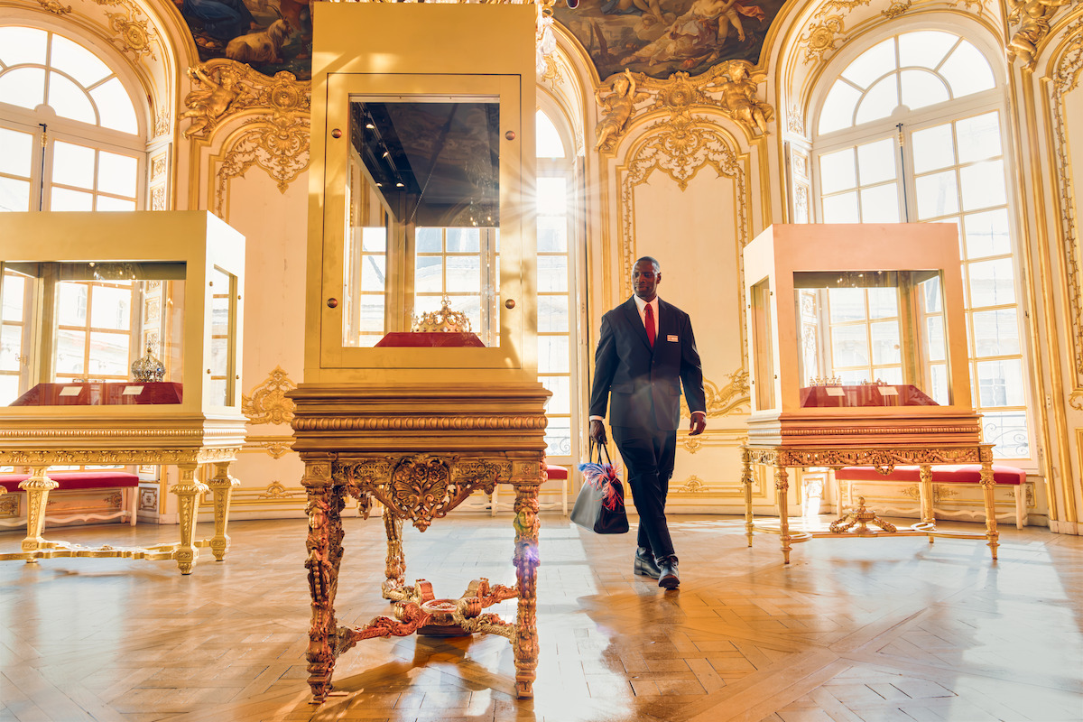 A security guard walks through an ornate, sunlit museum gallery with large windows, golden decorative walls, and glass display cases showcasing artifacts on elegant tables.
