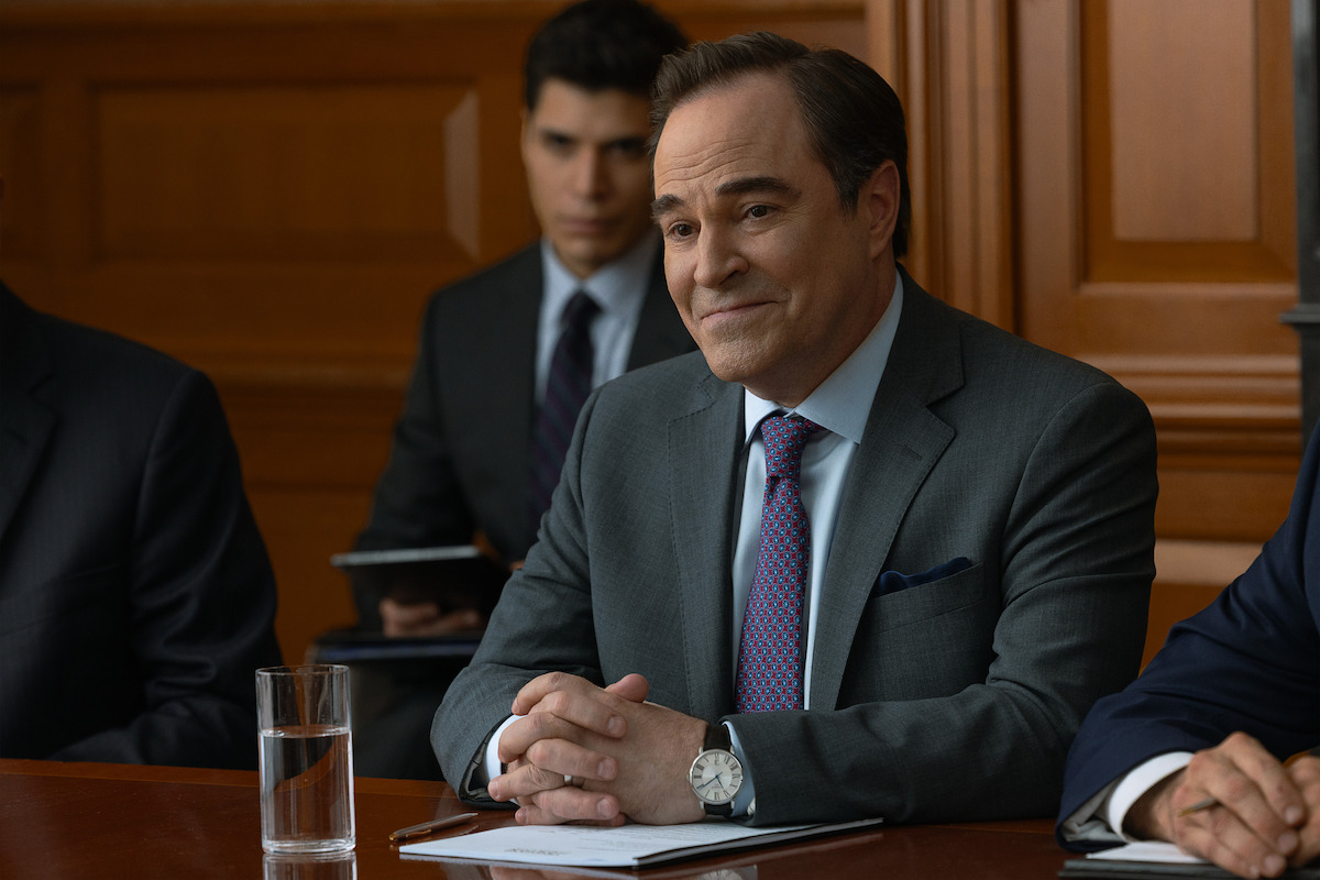 A man in a suit sits at a table in a courtroom or formal meeting room, with a glass of water, documents, and others around him, suggesting a legal or business setting. Wood paneling and another suited person are in the background.