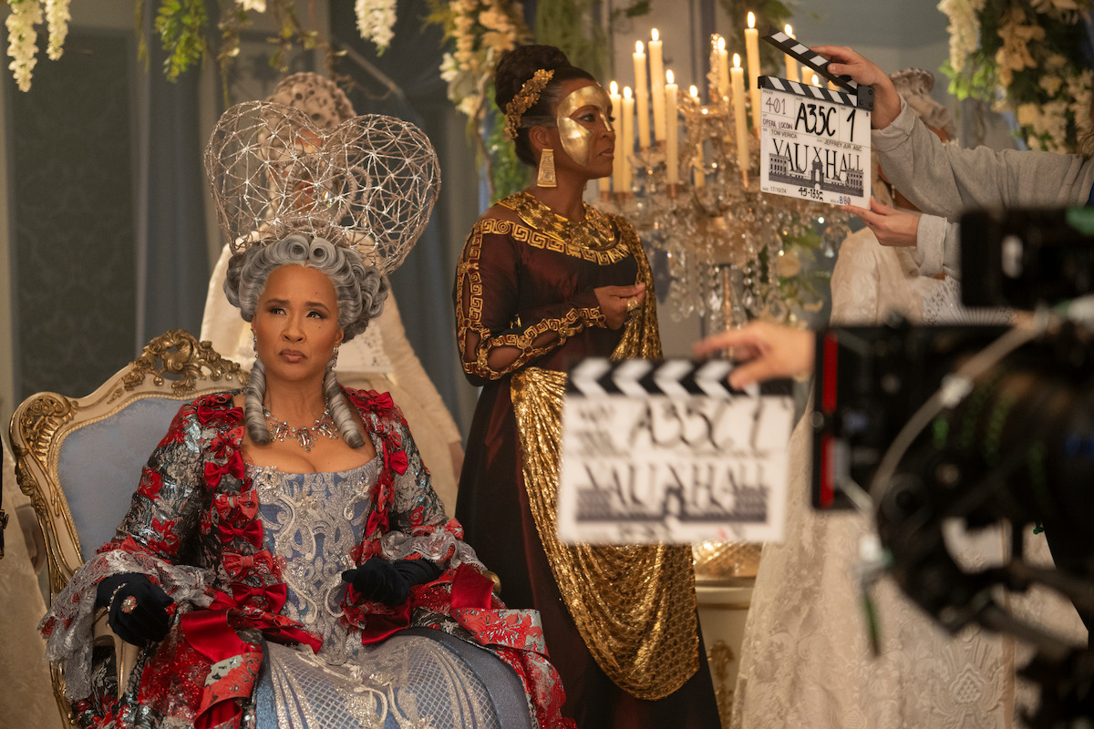 Woman in elaborate period costume sits on ornate chair; another woman in gold costume and mask stands behind her. Chandelier and flowers decorate the luxurious set while crew members hold clapboards, indicating a film shoot.