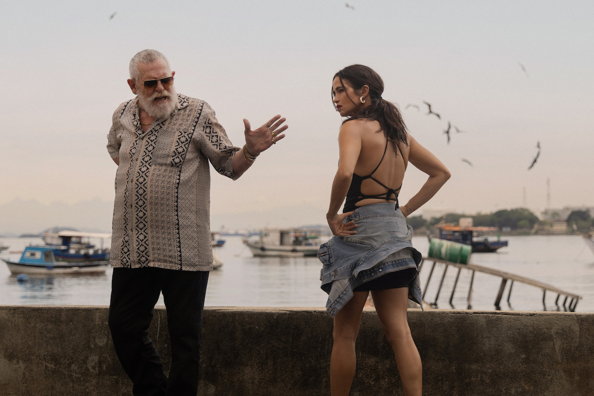 Um homem e uma mulher aparecem em uma praia com barcos de pesca e pássaros sobrevoando a água