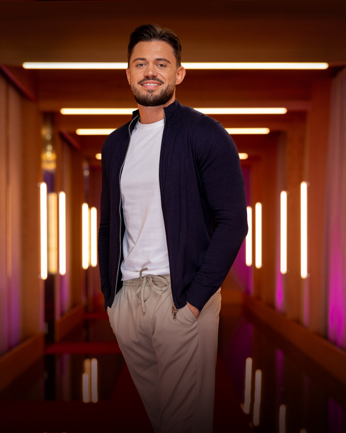 Man with dark hair and beard wearing a navy jacket and white shirt, standing in a modern, warmly lit hallway with vertical light fixtures and wooden accents, posing confidently with hands in pockets.
