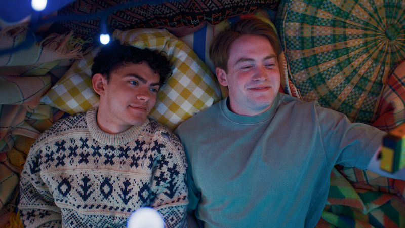 Two young men lying on colorful patterned blankets, one in a knit sweater and the other in a blue shirt, surrounded by pillows and soft lighting, creating a cozy, intimate indoor setting.
