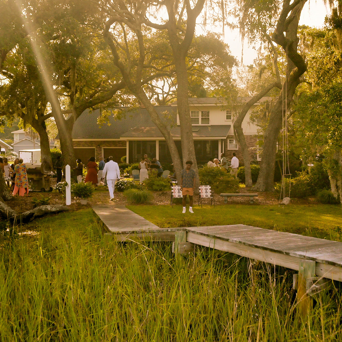 Pessoas reunidas no jardim de uma casa, cercadas de árvores grandes e vegetação, com a luz do sol entrando. Um píer se ergue do pântano rumo à entrada, evocando um evento tranquilo ou reunião de família em um ambiente natural ao ar livre.