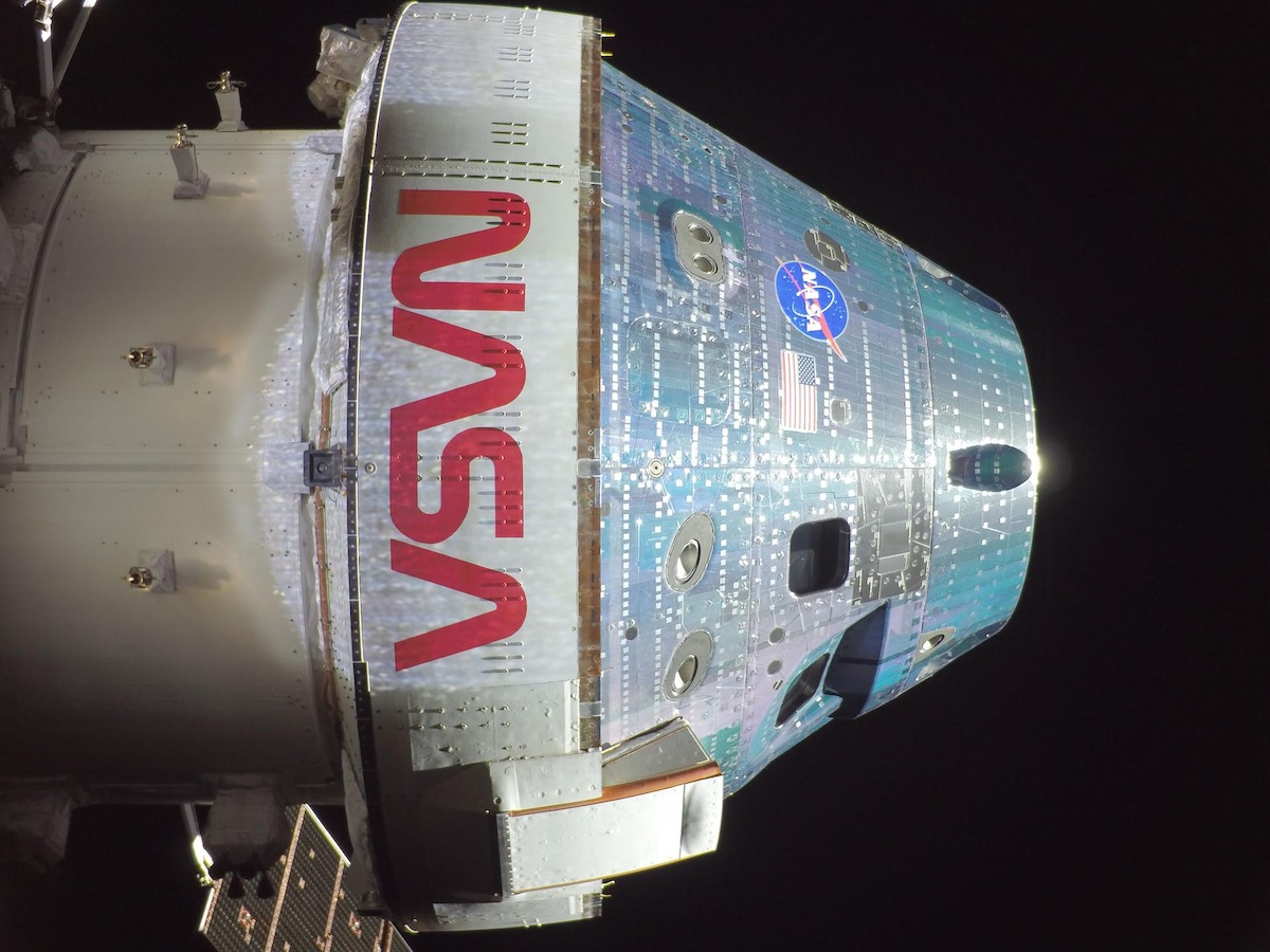 Close-up of the NASA Orion spacecraft in space, displaying its heat-shield exterior, NASA logo, American flag, and docking hatch under dark sky.
