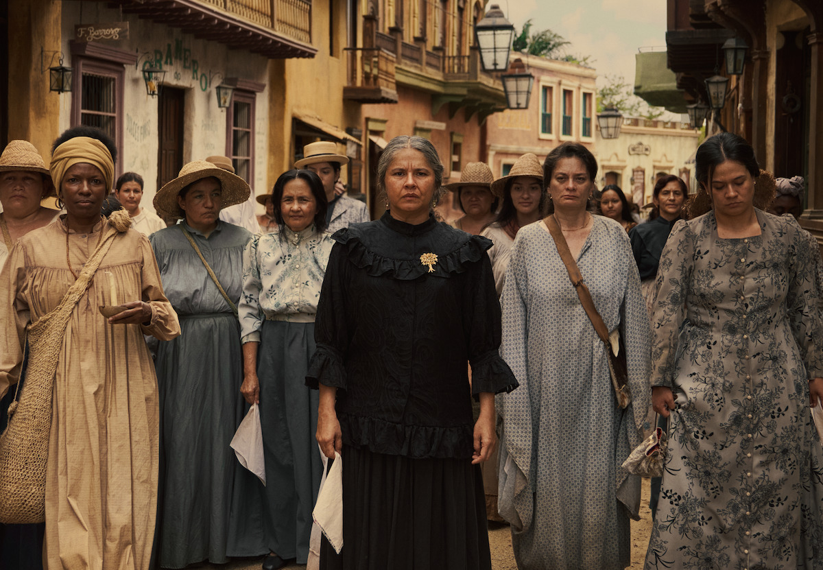 Group of women in historical clothing walking down a cobblestone street in a colonial town, looking forward with serious expressions, holding white cloths, surrounded by colorful old buildings and lanterns.