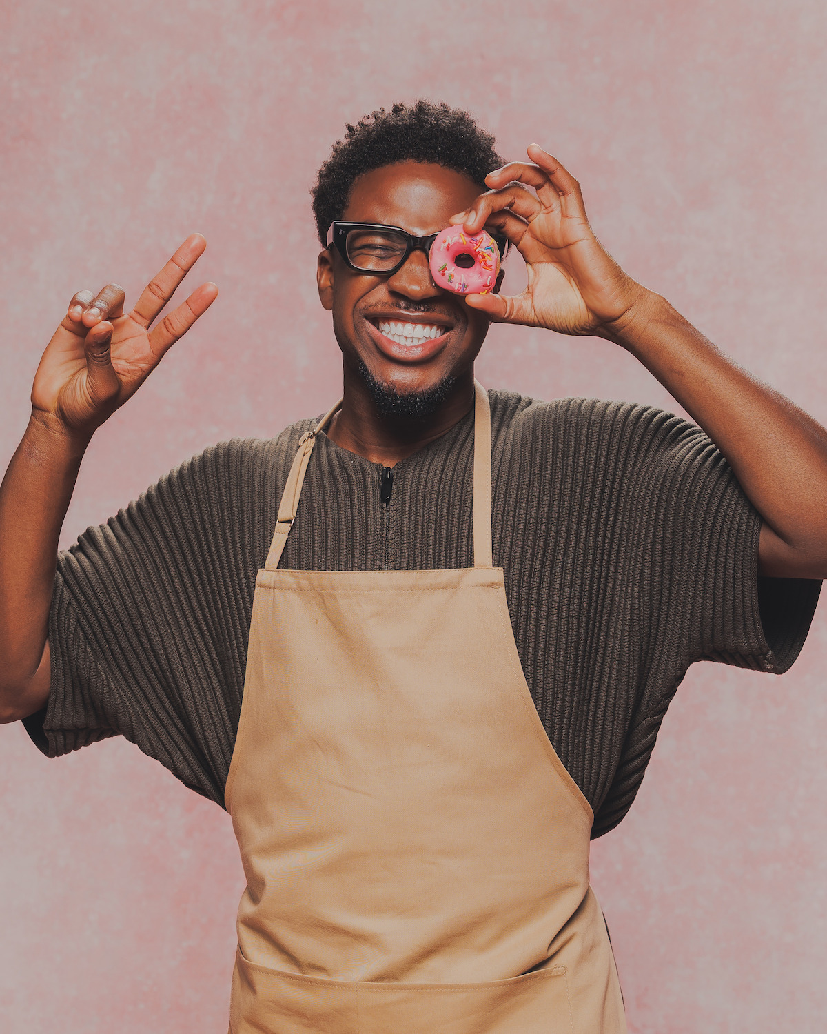 Smiling person in apron holding a pink donut up to their eye, making a peace sign with other hand, standing in front of a pink textured background.