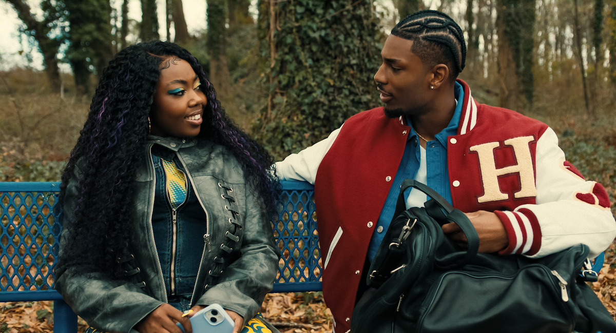 Two young people sit on a blue bench outdoors in a wooded park. The woman has long curly hair and bright makeup, holding a phone, while the man in a red varsity jacket talks to her, holding a black bag.