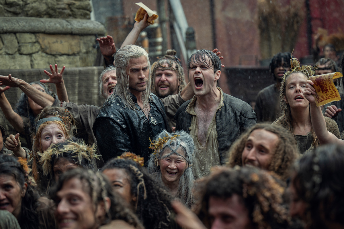 Two men, one with white hair and the other shouting, stand amid a lively, muddy medieval crowd celebrating outdoors. The mood is intense and chaotic, with expressions of joy and excitement.