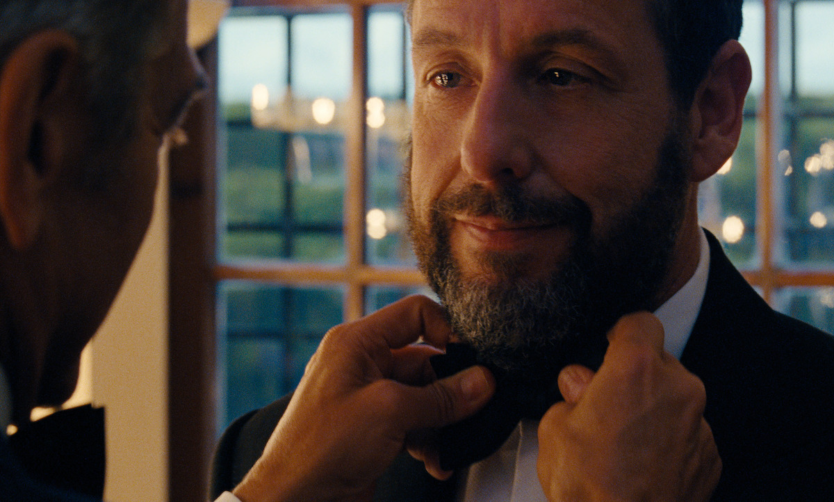 A man in formal attire has his bow tie adjusted by another person in a warmly lit room with large windows, creating a calm, intimate, and elegant atmosphere.