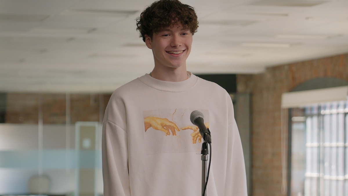 Smiling young person stands at a microphone in a modern, bright office with brick walls, wearing a white sweatshirt with a Renaissance-style hands print. The mood is cheerful and relaxed.