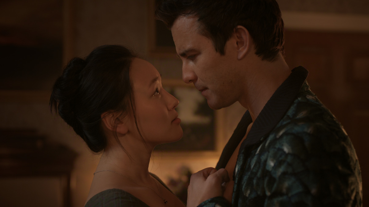 A woman and man stand close together in a dimly lit, cozy room, gazing into each other's eyes with intense expressions, suggesting a moment of romantic tension or emotional connection.