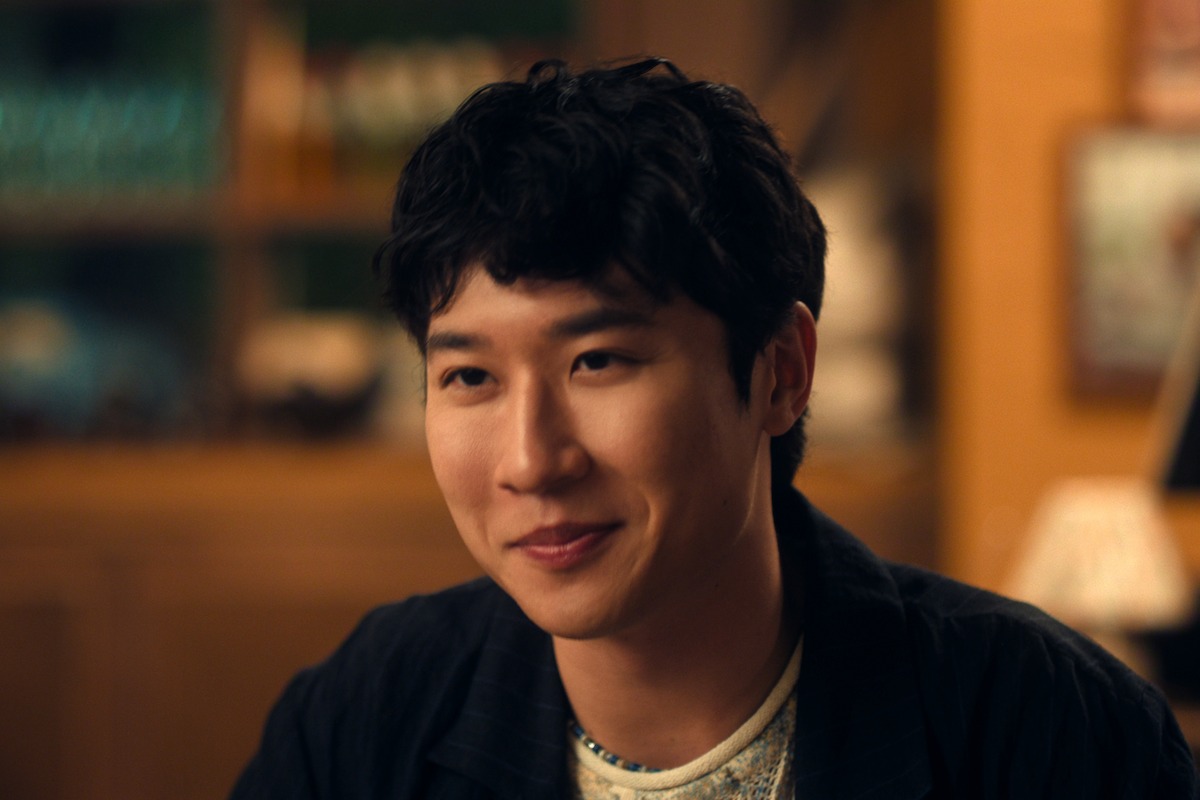 A young man with dark hair smiling indoors, sitting in a warmly lit room with blurred shelves and decorations in the background, suggesting a cozy home or café atmosphere.