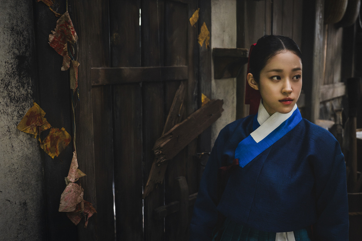 Woman in traditional Korean hanbok standing near an old wooden door with dried leaves, looking to the side in a dimly lit rustic setting.