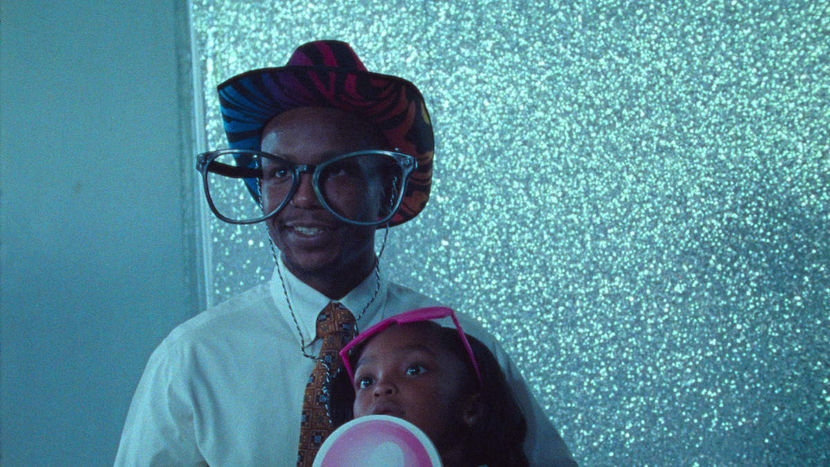 Aubrey Smith sits on Keith Sweptson's lap. The latter wears oversized sunglasses and a cowboy hat.