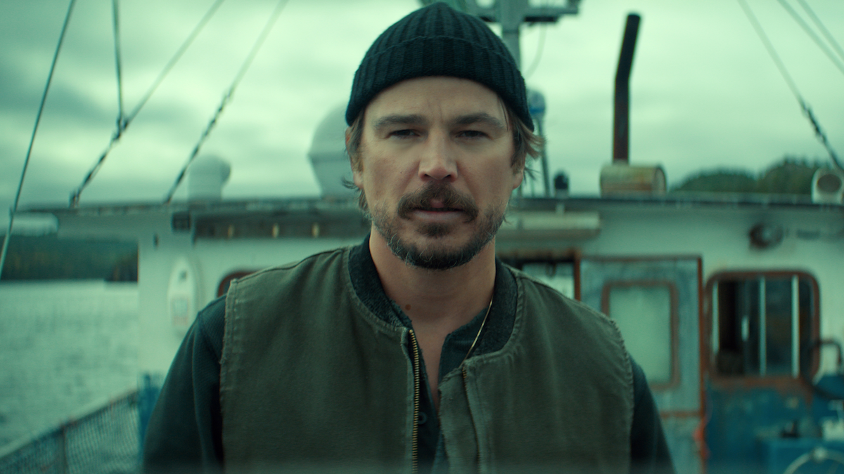 A man wearing a black beanie and green vest stands on a fishing boat, with water and cloudy sky in the background, conveying a rugged, maritime setting.