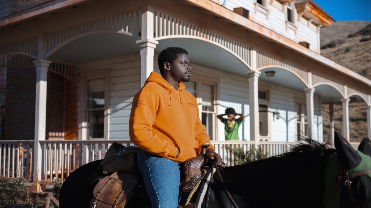 Man in orange hoodie sitting on a horse in front of a large white house with arches, sunny day, another person standing on porch in the background.