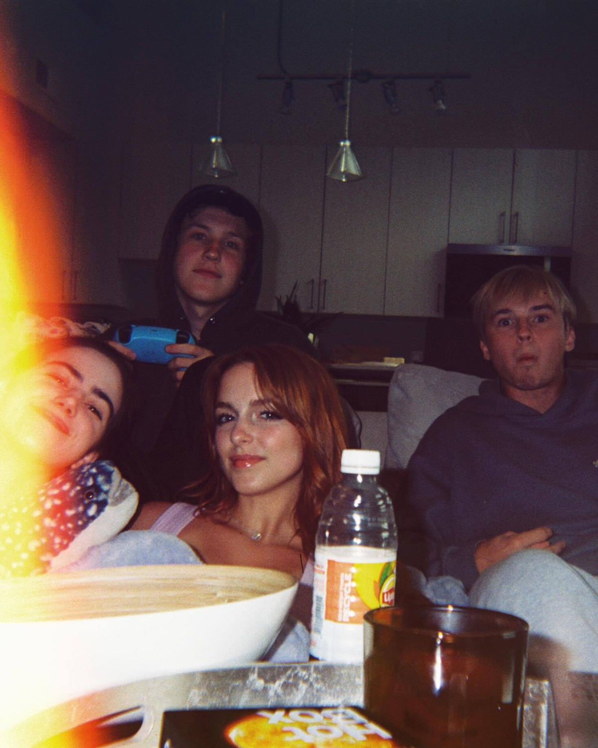 Four friends sit on a couch in a cozy living room or apartment kitchen, relaxing with drinks, snacks, and a video game controller visible; a light leak is present on the left side of the image.