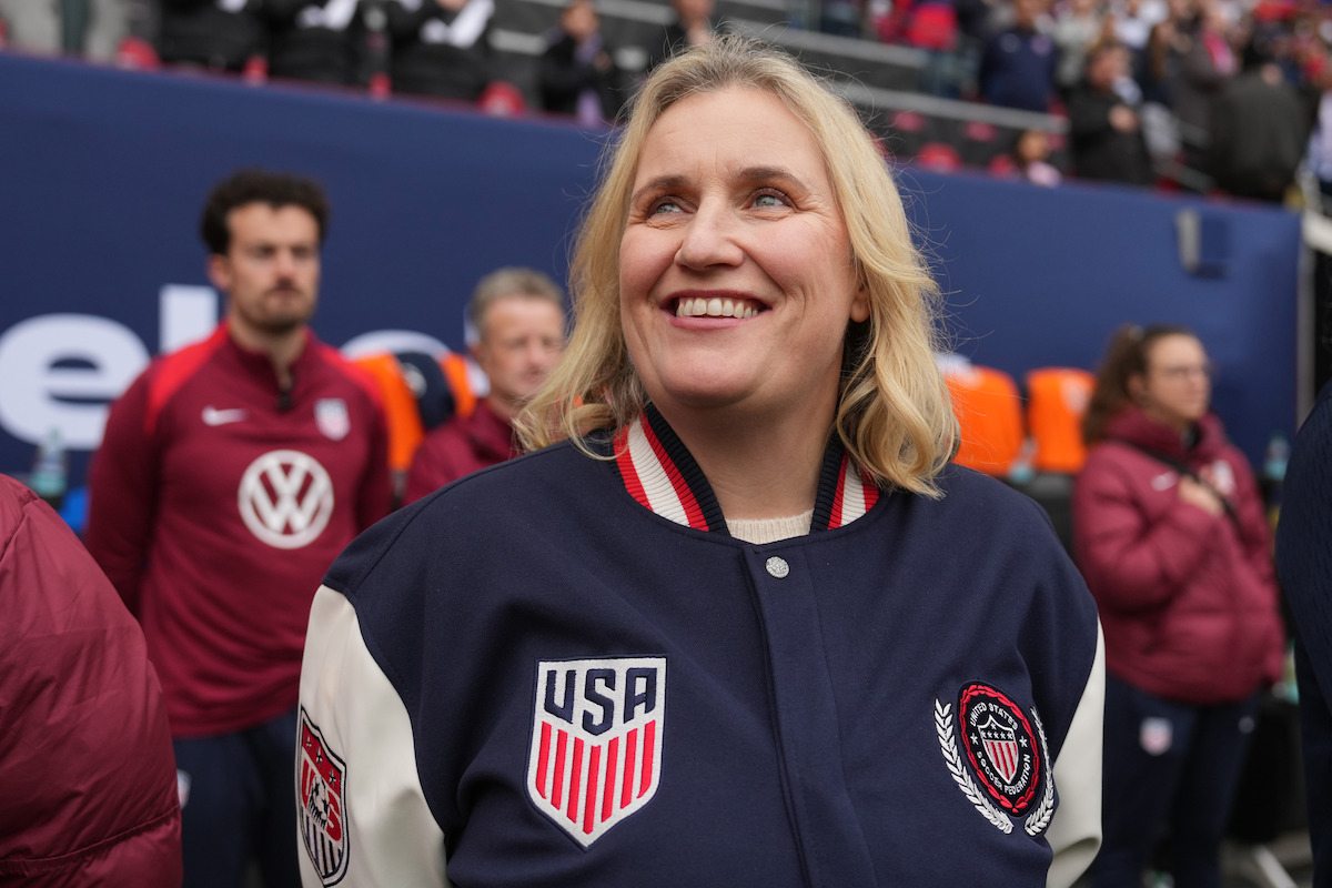Smiling woman in a USA soccer jacket stands on a stadium sideline with blurred teammates and crowd in the background, appearing happy and engaged before a sports event.