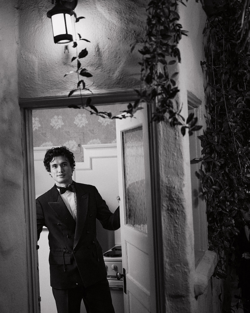 A man in a suit stands in a doorway, partially inside a house with patterned wallpaper, under a lit outdoor lantern, and surrounded by climbing plants in a black-and-white setting.