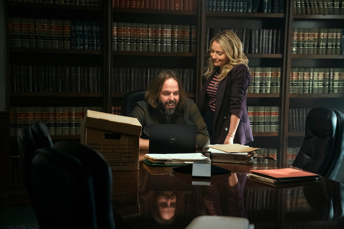 A man with long hair sits at a desk as a woman with blonde hair stands behind him.