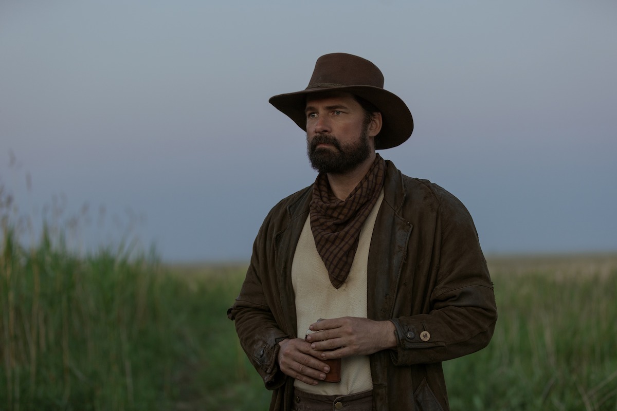 Man dressed as a cowboy with a brown hat and bandana stands in a grassy field at dusk, looking into the distance. The sky is clear and the atmosphere is calm, evoking a western or frontier setting.