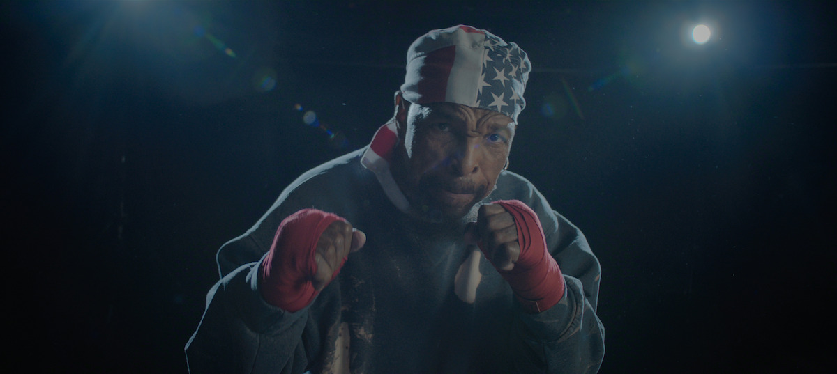 A boxer wearing red gloves and an American flag bandana is posing in a fighting stance in a dark, dramatic setting with bright lights in the background.