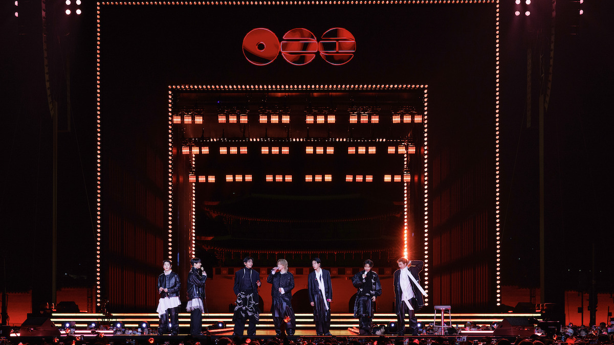 SUGA, JIMIN, JIN, JUNG KOOK, V, J-HOPE, and RM on a brightly lit stage with orange and yellow lights, geometric decorations, and an audience watching a night concert.