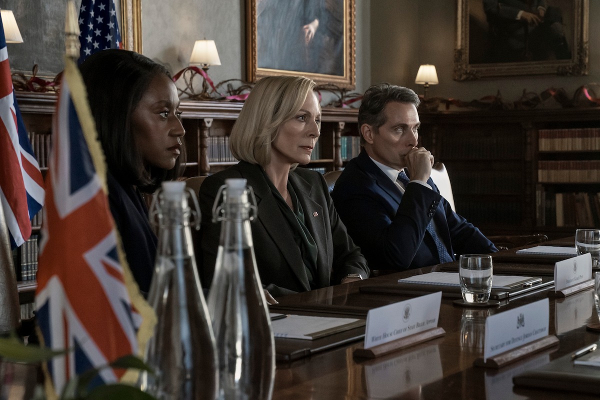 Nana Mensahas as Billie Appiah, Allison Janney as Grace Penn, and Rufus Sewell as Hal Wyler sit at a government meeting while they listen quietly.