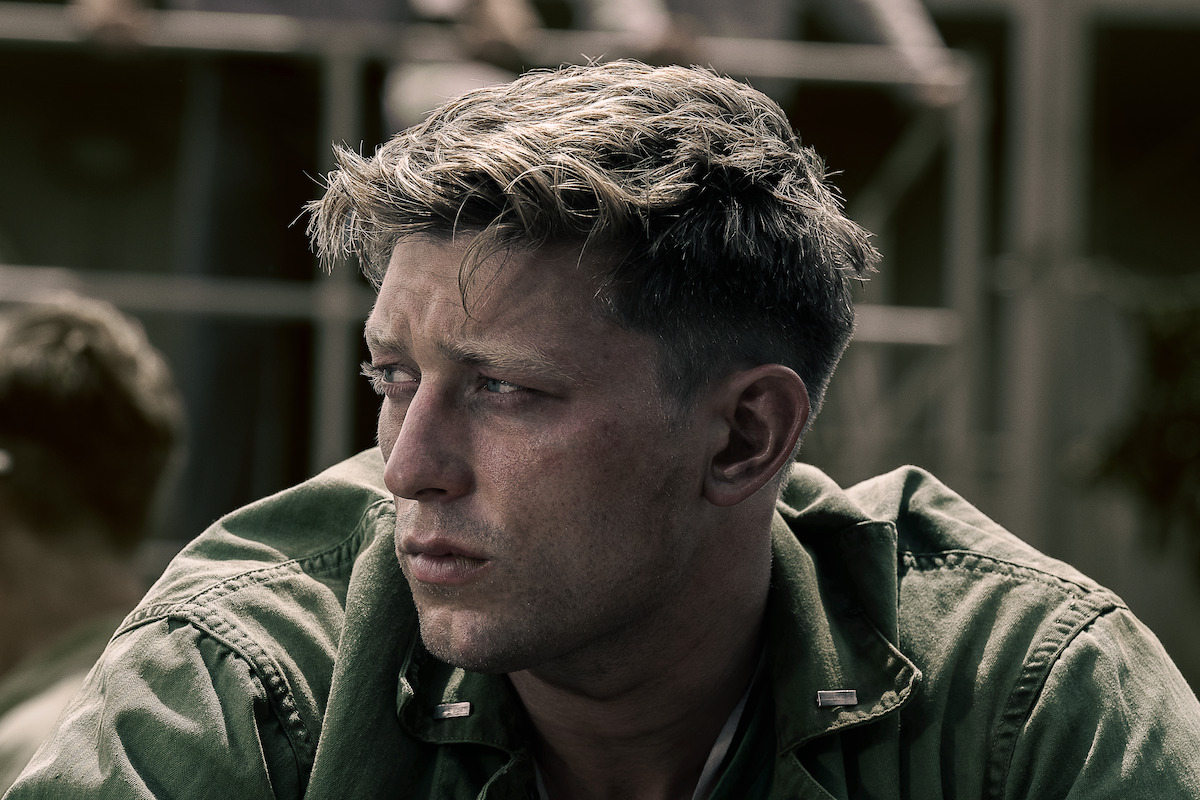 Tom Francis as Dave Schreiner in military uniform with short hair looks to the side with a serious expression, outdoors with blurred structures and another person in the background, suggesting a wartime or military setting.