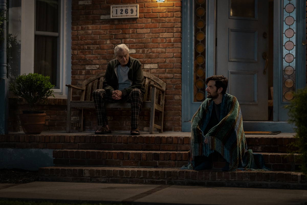 Two men sit outside a brick house at night; one on a bench looking down, the other wrapped in a blanket on the porch steps, suggesting a quiet, contemplative moment. The porch is dimly lit, evoking a calm, reflective mood.