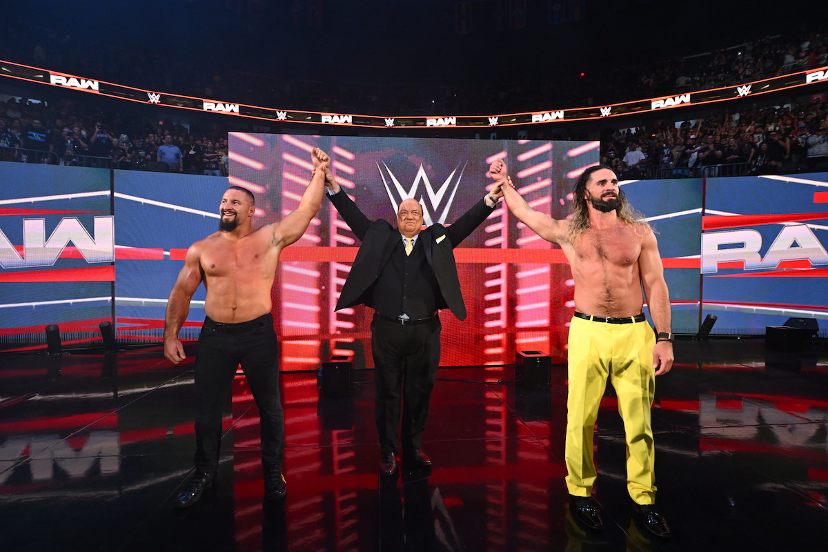 Bron Breakker, Paul Heyman, and Seth Rollins holding hands raised in victory. Bright arena lights and a cheering crowd are visible in the background.