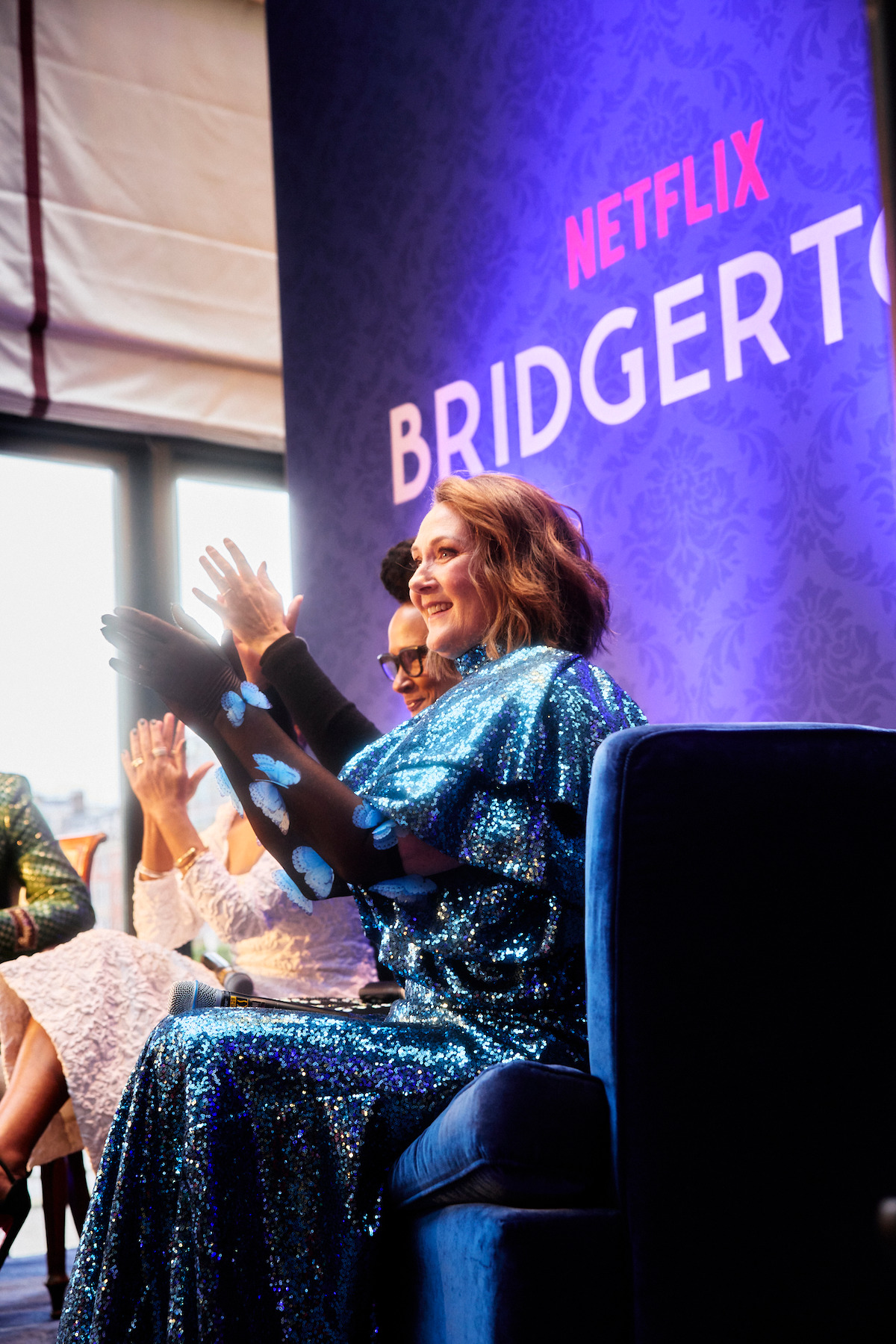 Woman in a blue sequin dress smiling and clapping at a Netflix Bridgerton event, seated indoors with others in a well-lit room with purple decor.