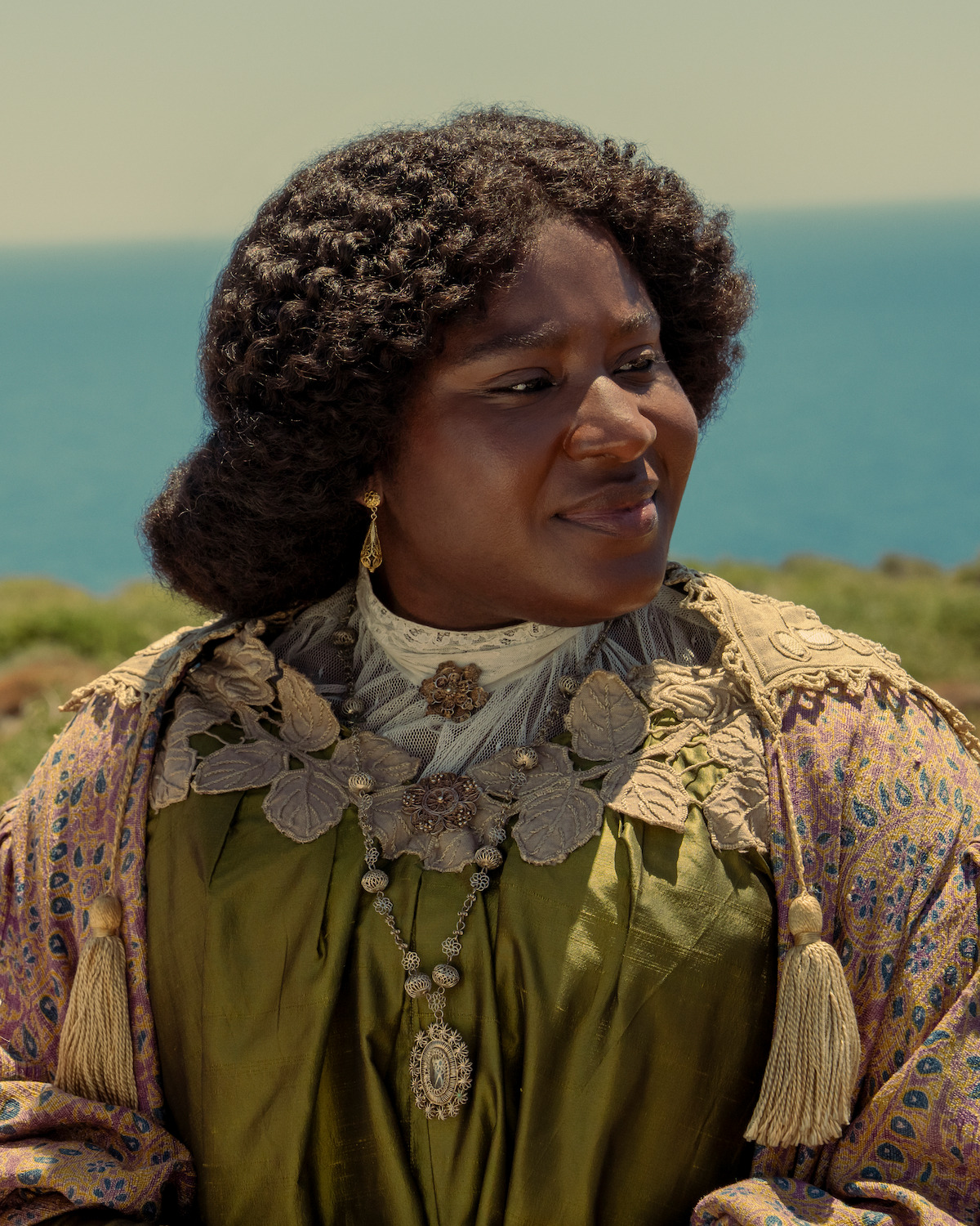Woman in ornate green dress with lace and jewelry stands outdoors near the sea, wearing historical clothing with intricate details. Sunny, coastal landscape with greenery and blue sky in the background.
