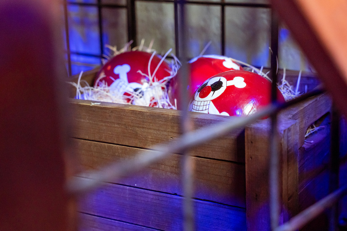 Red eggs with skull and bone designs sit in straw inside a wooden crate, surrounded by a metal cage, with blue and purple lighting creating a mysterious atmosphere.