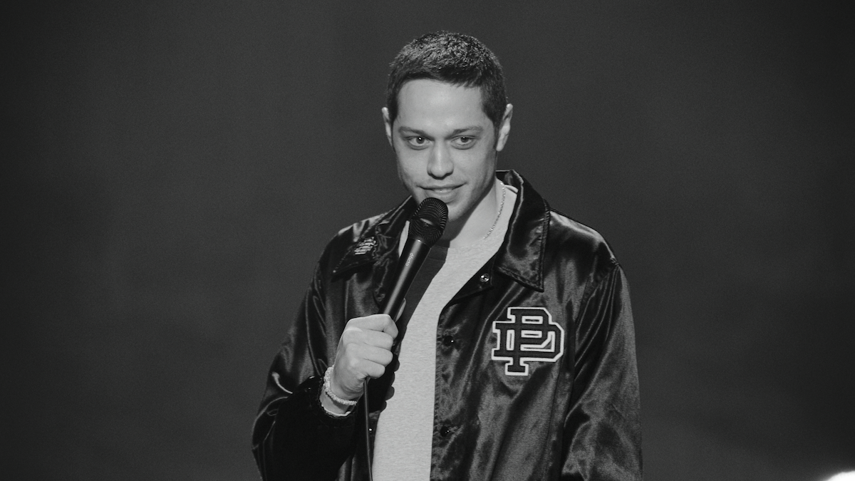 A person in a satin jacket stands onstage holding a microphone, performing stand-up comedy in front of a dark, blurred background. The image is in black and white.