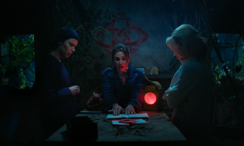 Three people gather around a dimly lit table with red symbols in a mysterious, overgrown room, discussing or examining papers, creating a tense and secretive mood. A strange diagram is drawn on the wall behind them.