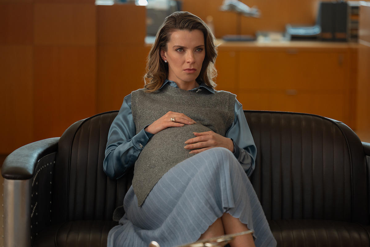 A pregnant woman sits on a black leather couch in an office setting, wearing a blue blouse and gray vest, looking serious and contemplative.