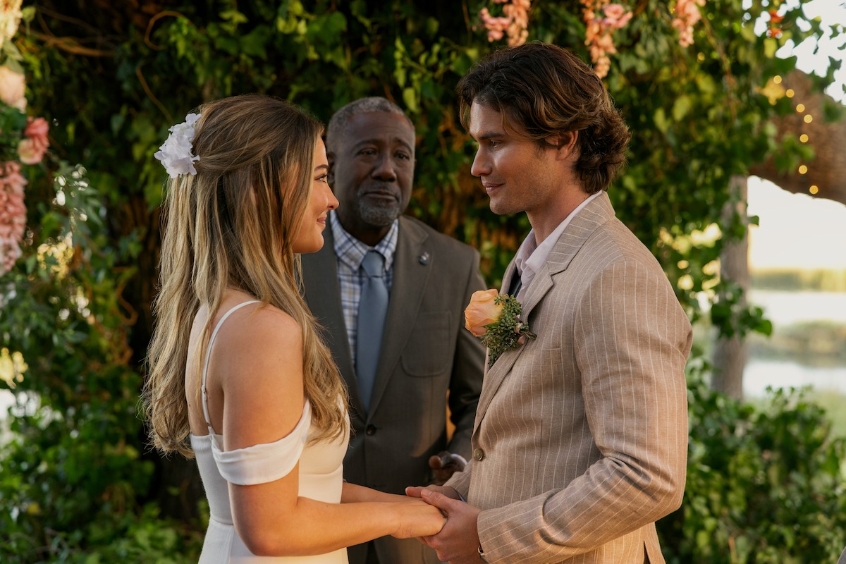 Bride and groom holding hands at an outdoor wedding ceremony, standing under a floral arch with an officiant present, surrounded by greenery and flowers.