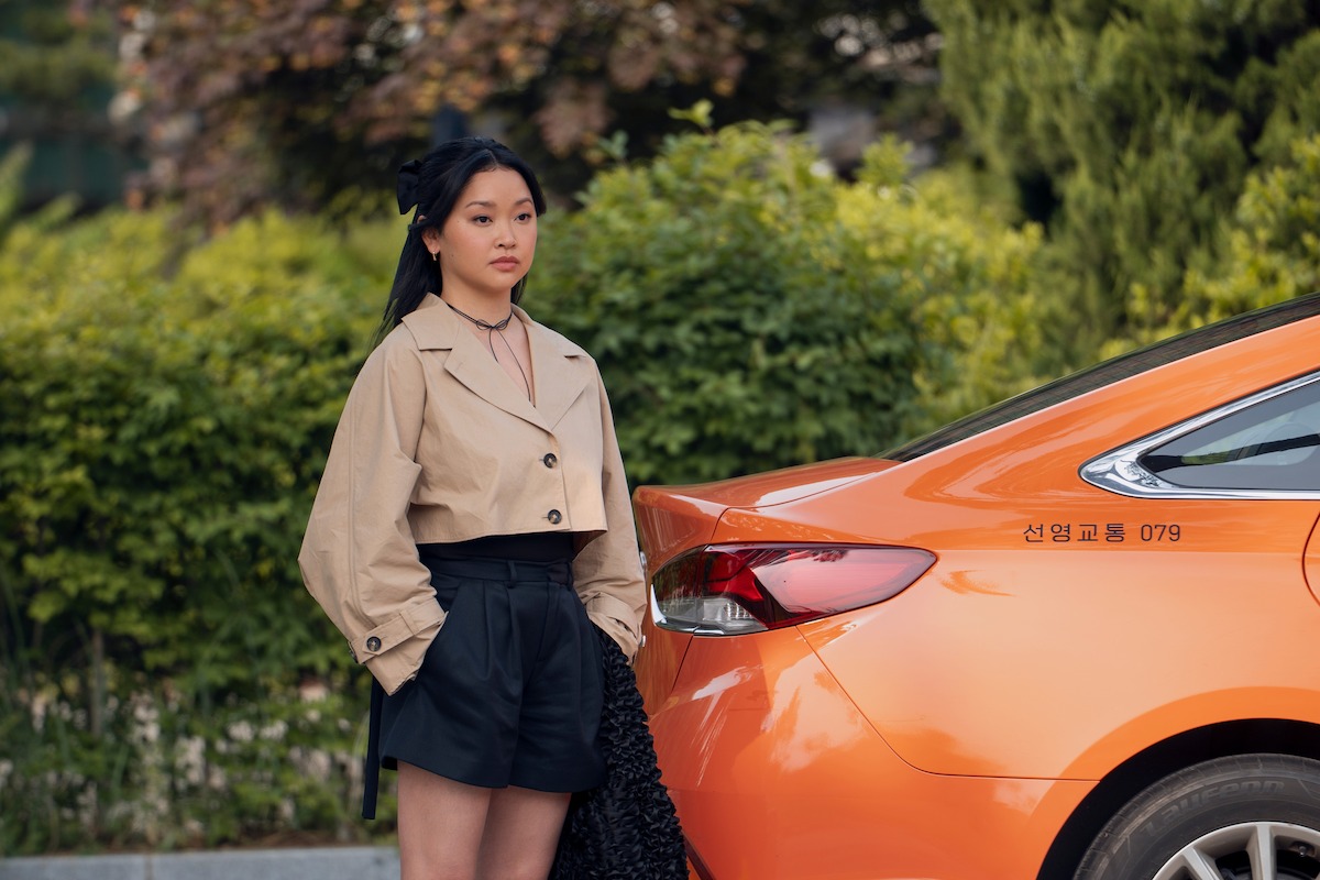 A woman in a beige jacket and black shorts stands next to an orange car with Korean writing, outdoors near green bushes and trees on a sunny day.