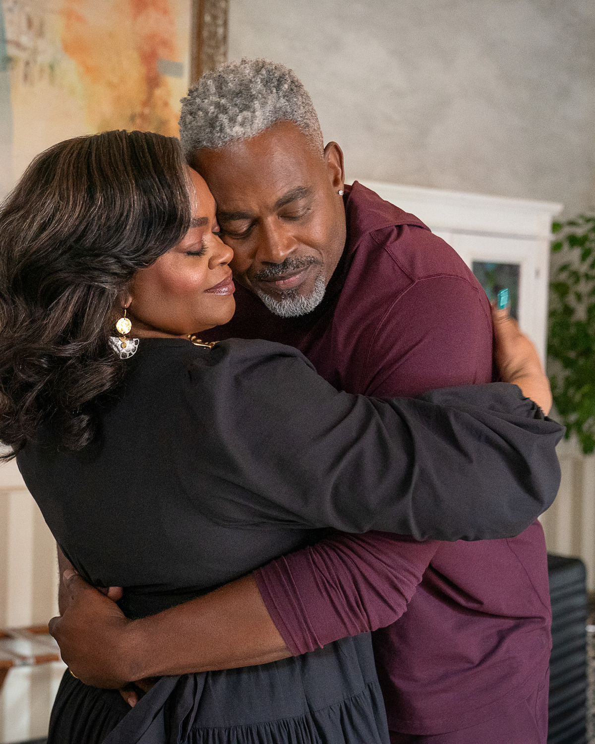 Middle-aged couple warmly embracing in a cozy, well-decorated living room with soft lighting and artwork in the background, conveying comfort and affection.