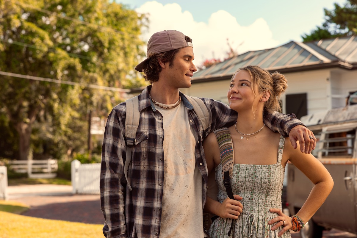 Young man with arm around young woman, both smiling and standing outdoors on a sunny day. They appear happy and relaxed in a casual suburban neighborhood, with trees, a house, and vintage van in the background.