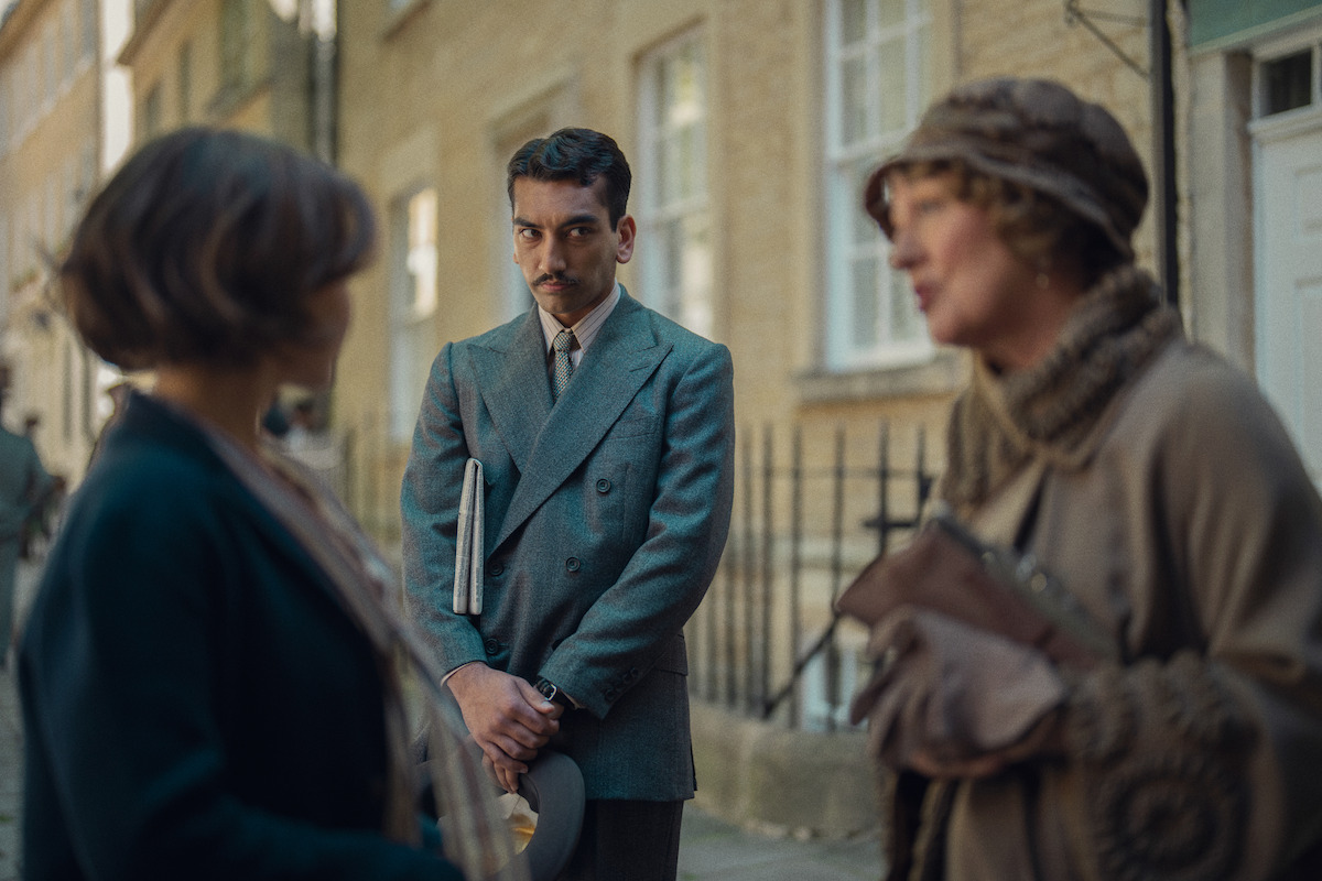 A man in a gray suit holding a notebook stands on a city street, looking at two women in conversation; the setting appears historical with people in period clothing and old brick buildings in the background.