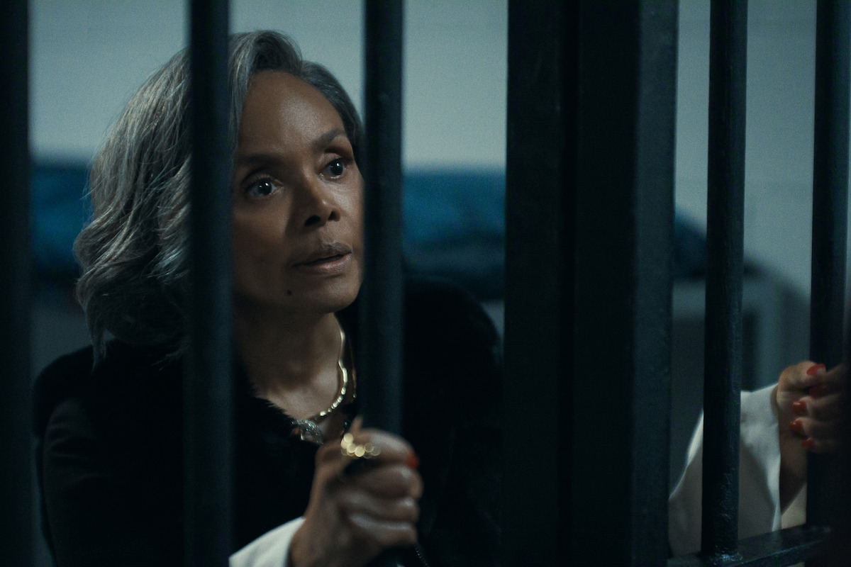 A woman with gray hair grasps jail bars in a dimly lit cell, looking intently outward. The environment suggests a prison or detention setting with a somber and tense atmosphere.