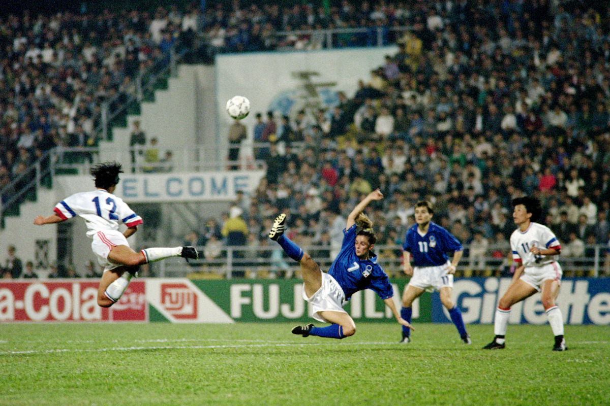Soccer match action with player in white and red jersey kicking the ball mid-air, blue team players defending, stadium crowd in background, night setting, energetic atmosphere.