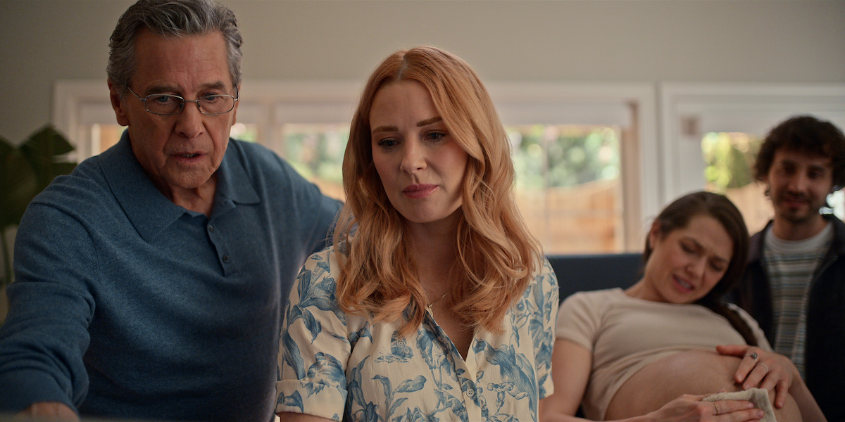 Four people indoors: an older man and a woman in front, while a pregnant woman lies on a couch with a supportive man beside her.