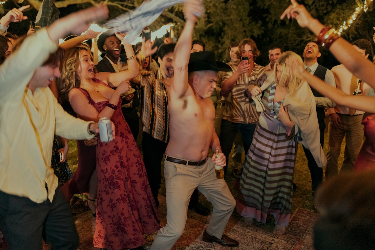 Shirtless man in cowboy hat dancing energetically at an outdoor party at night, surrounded by cheering people in festive attire with string lights and trees in the background.